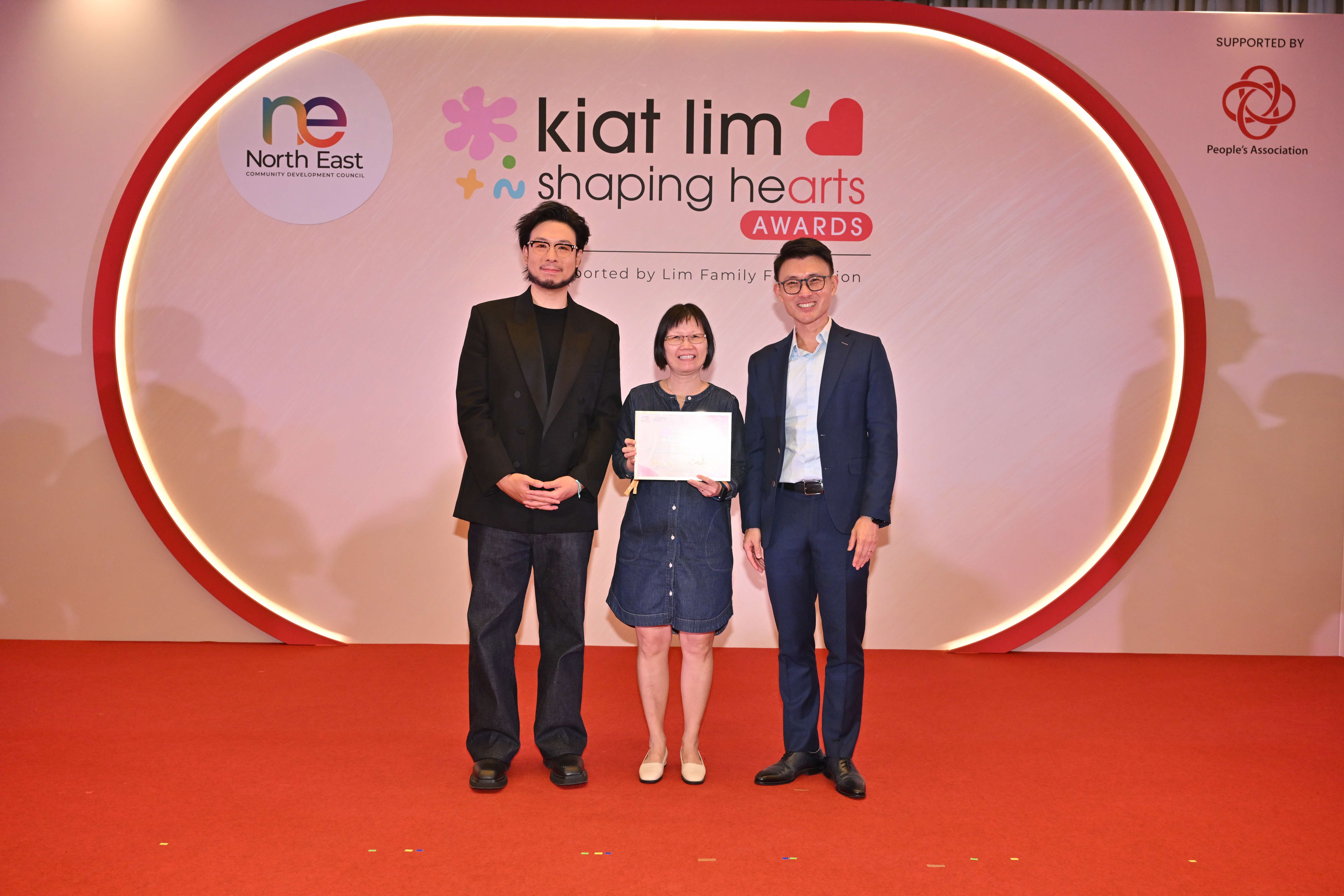 Award recipient standing on stage with the presenter, holding their certificate and posing for a group photo during the award ceremony, with a backdrop reading ‘Kiat Lim-Shaping Hearts Awards'.